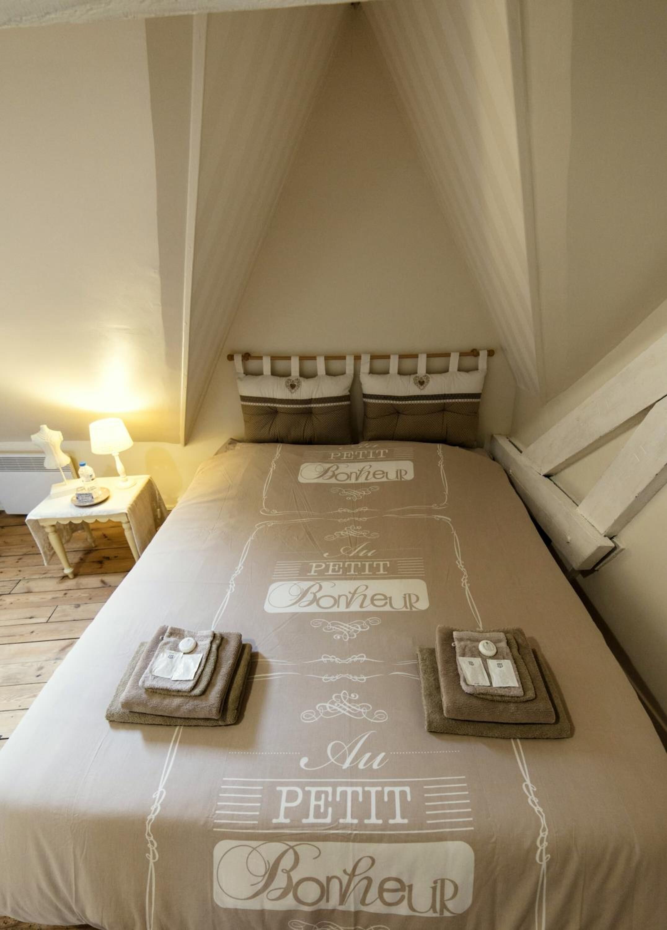 Een slaapkamer met een hemelbed, een nachtkastje met een lamp, en een muur versierd met kussens en een tekst.