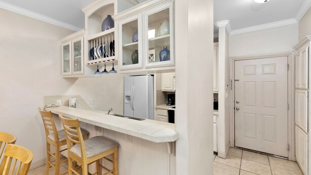 A compact kitchen with a white countertop, wooden chairs, and cabinets with glass doors displaying dishes.