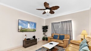 A living room with a sofa, a coffee table, a TV mounted on the wall, a ceiling fan, and a window with curtains.