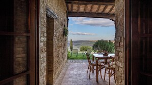 Une porte ouverte donne sur un patio aménagé avec une table et des chaises, d’où l’on peut admirer un paysage pittoresque composé de collines et d’arbres.