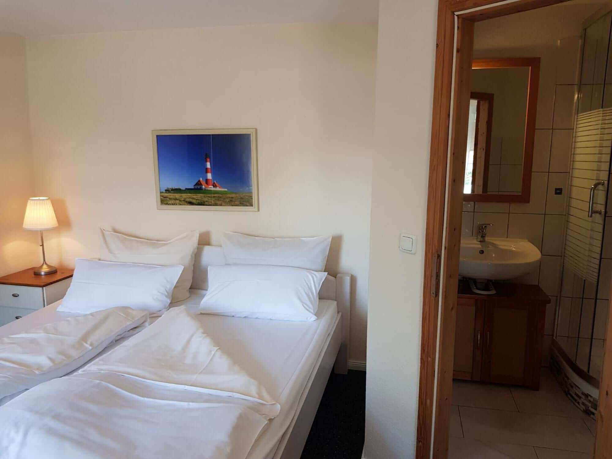 A hotel room with a bed, a nightstand with a lamp, a painting of a lighthouse, and a bathroom with a sink and mirror.