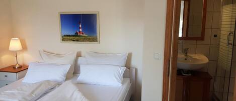 A hotel room with a bed, a nightstand with a lamp, a painting of a lighthouse, and a bathroom with a sink and mirror.