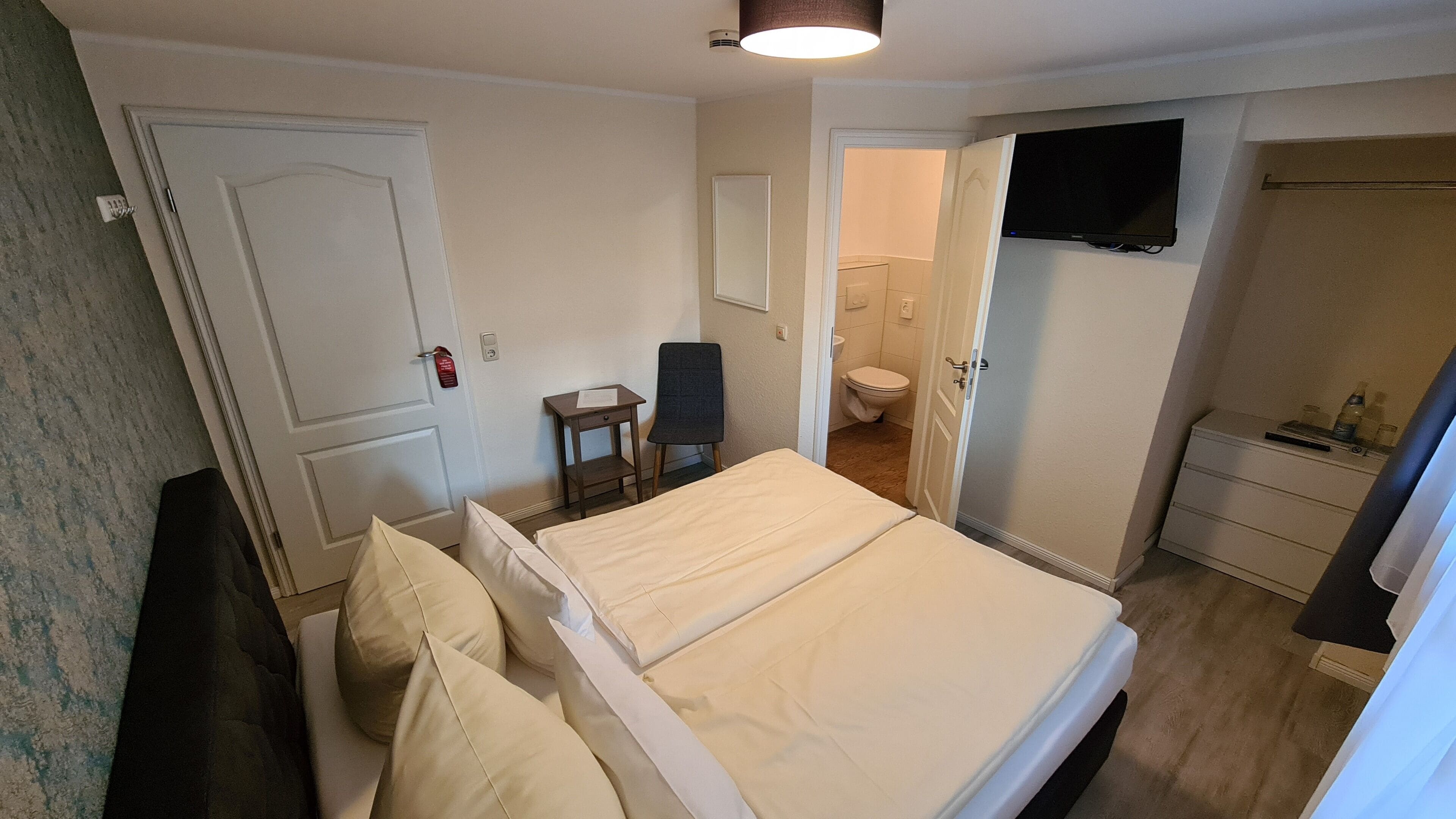 Quarto de hotel com cama, cadeira, escrivaninha, televisão e banheiro visível por uma porta aberta.