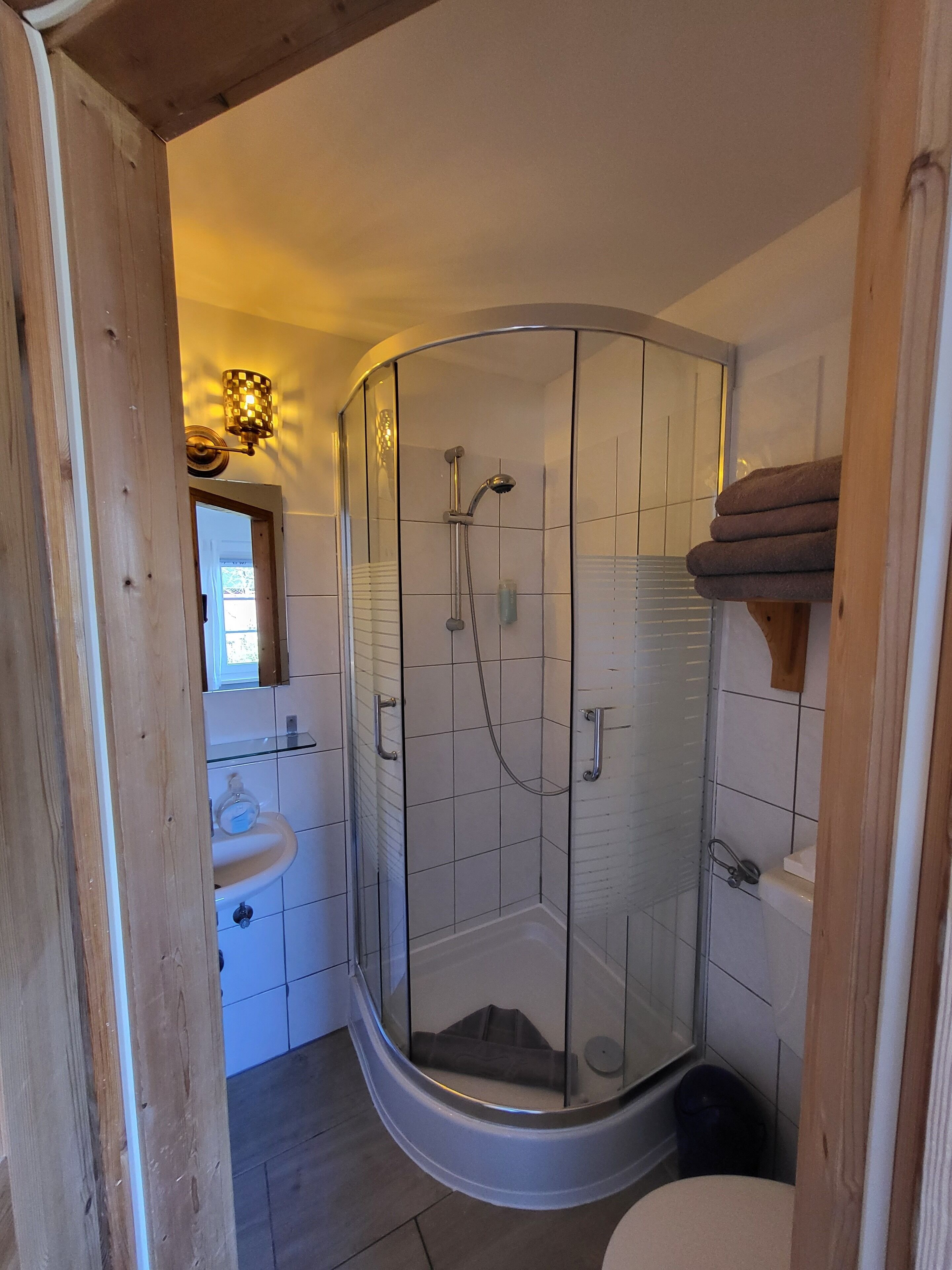 Ducha de esquina con puertas de vidrio, un lavamanos y un inodoro en un baño con vigas de madera y azulejos blancos.