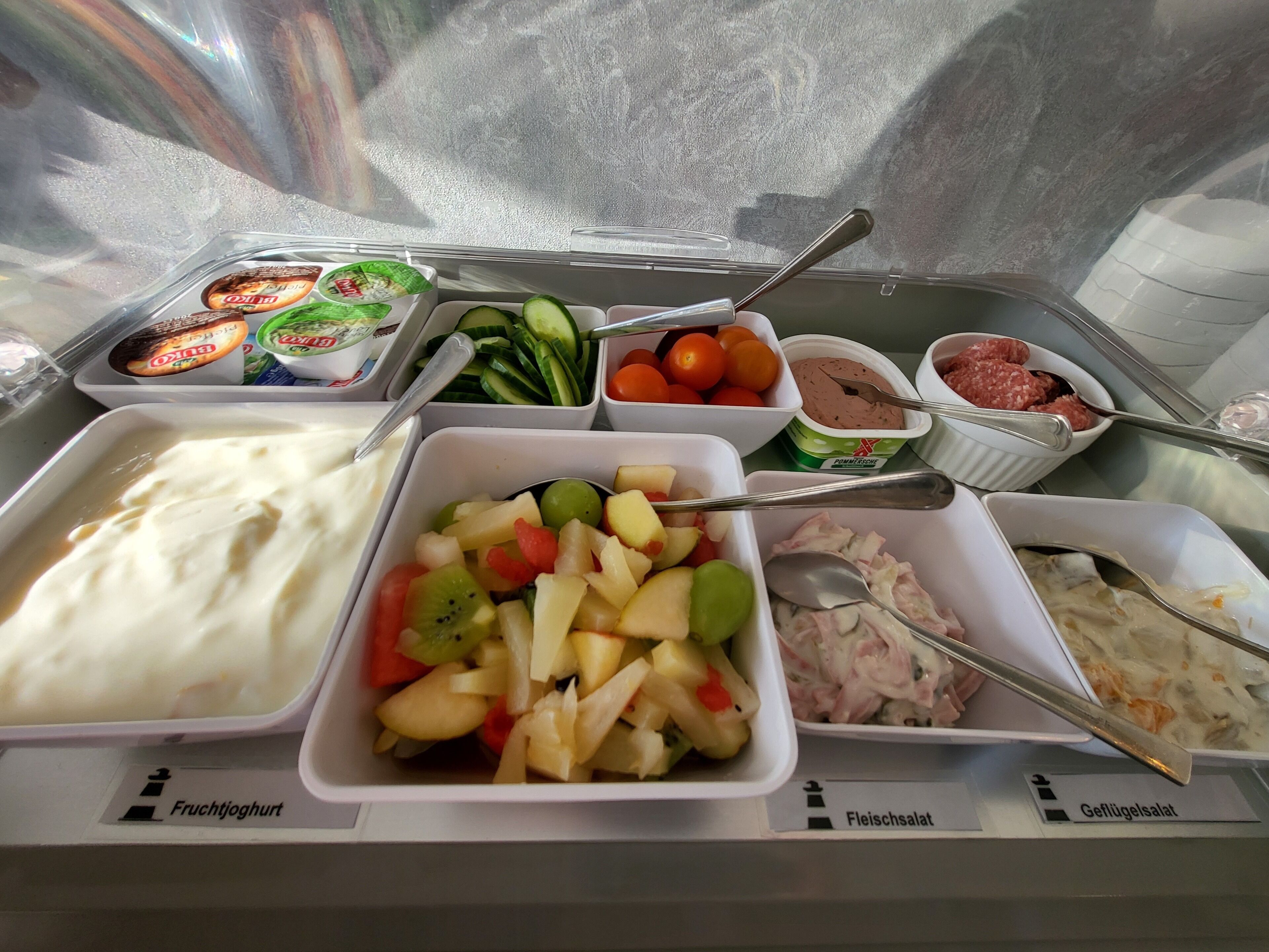 Een buffet met aangeduide gerechten, waaronder fruit yoghurt, komkommers, tomaten, vleessalade en een fruitsalade.