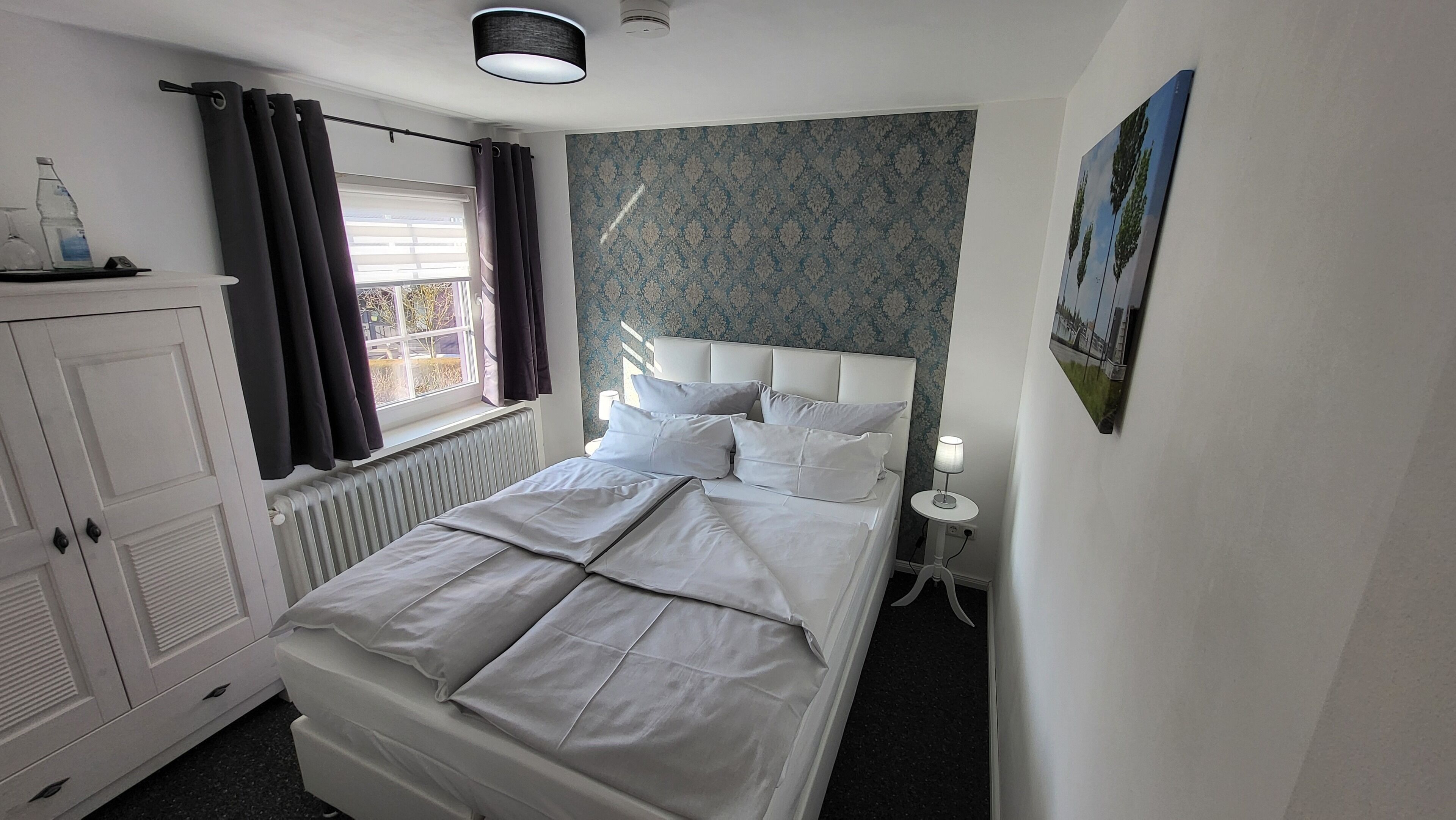 Un dormitorio ordenado con cama, radiador, ventana con cortinas, lámpara de pared y un cuadro.
