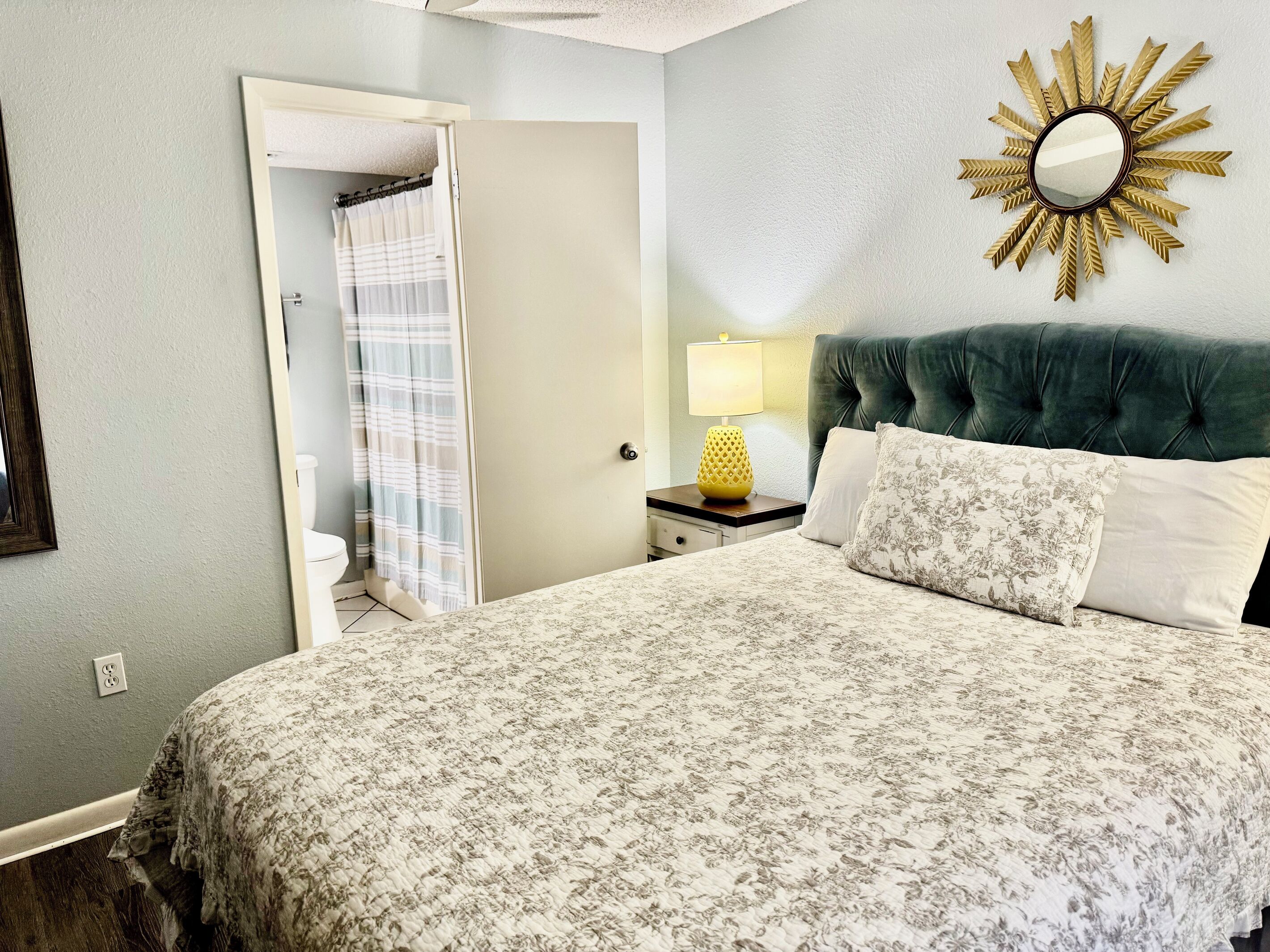 A bedroom with a bed, a nightstand with a lamp, a mirror, and a door leading to a bathroom.