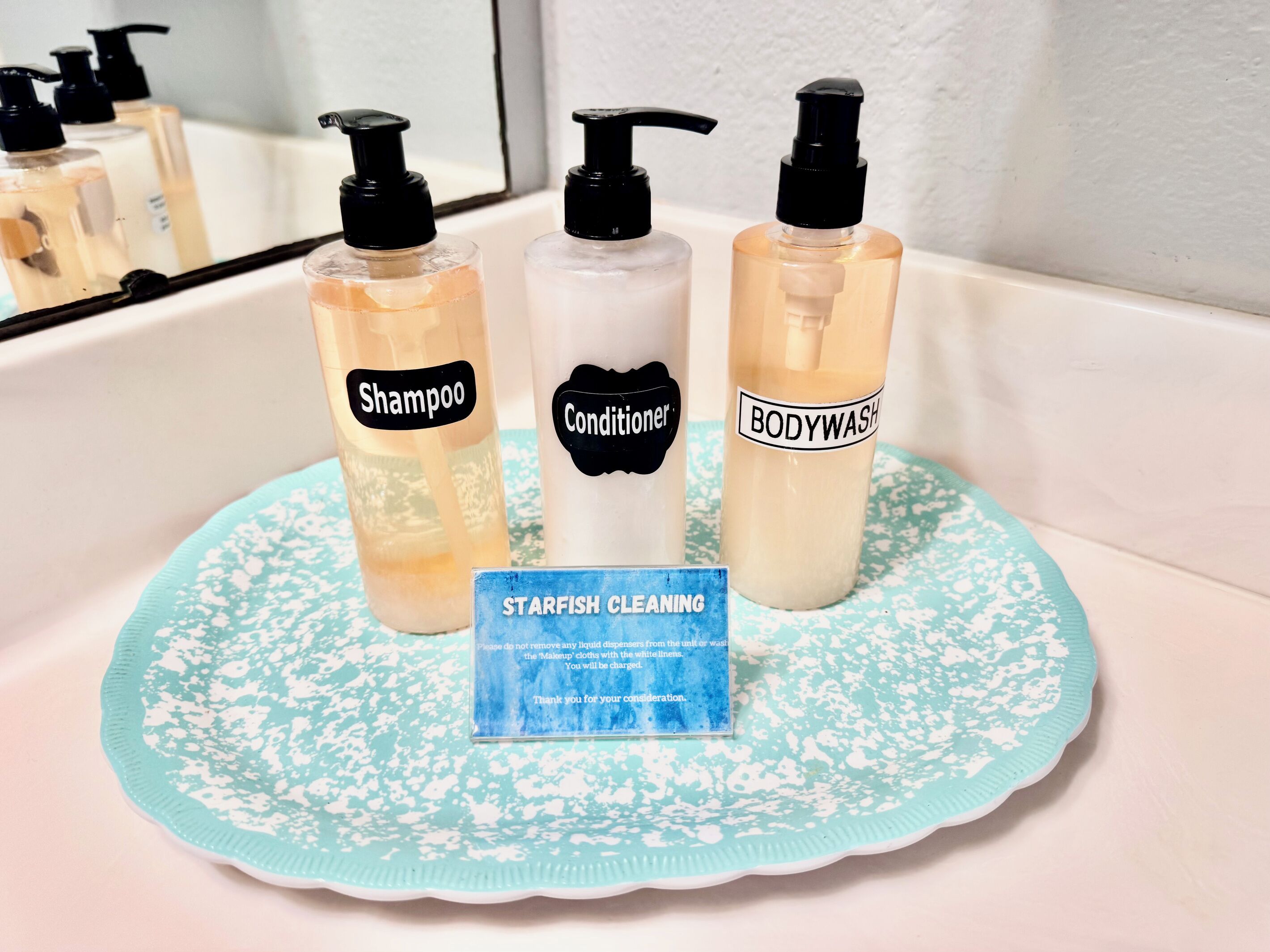 En håndvask med tre flasker personlig pleje med påskrifterne 'Shampoo', 'Conditioner' og 'Body Wash'. Et blåt rengøringsinstruktionskort ligger på håndvasken.