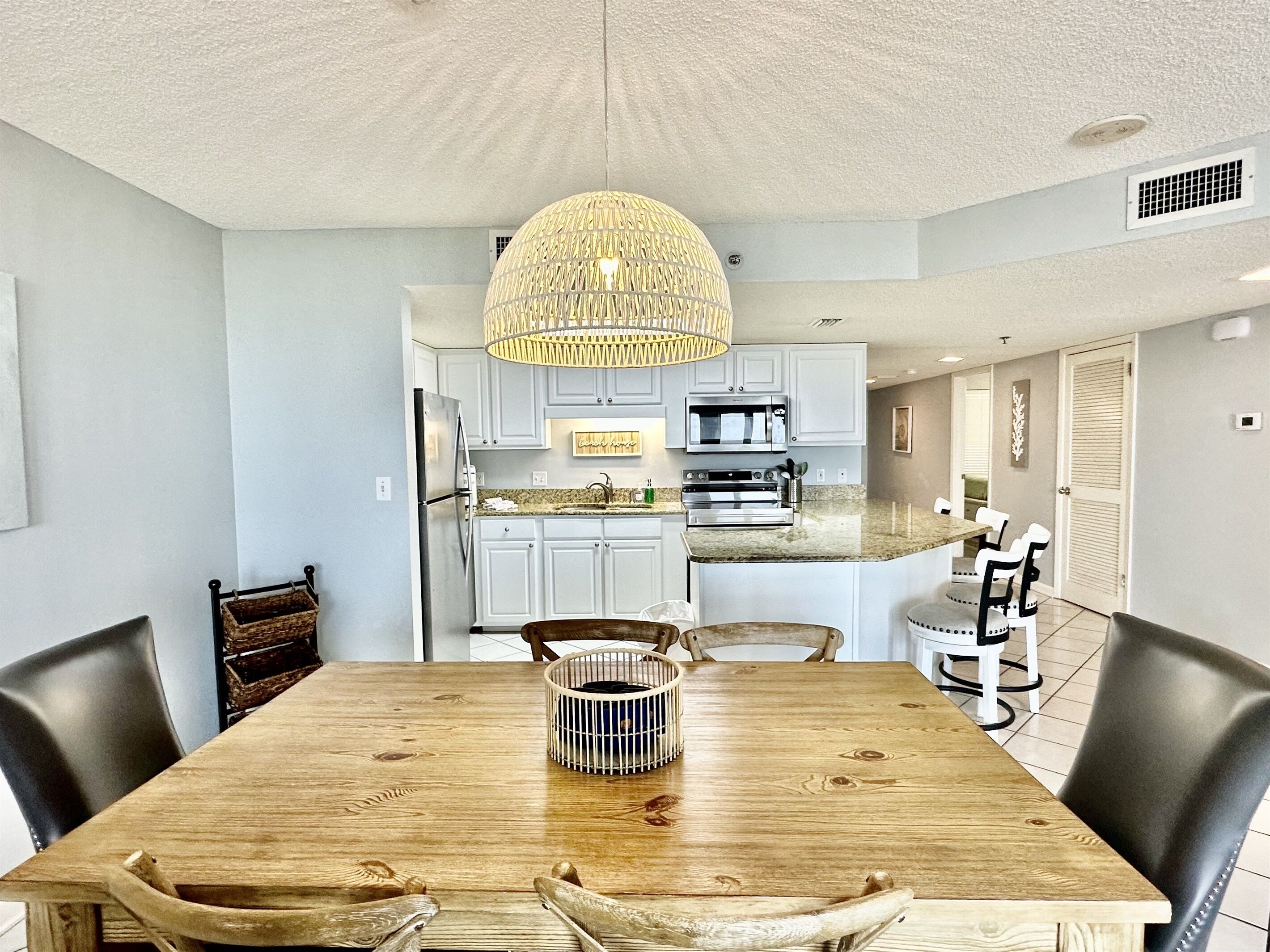 Een eetgedeelte met een houten tafel, stoelen, een geweven hanglamp en een keuken op de achtergrond.