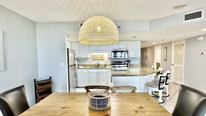 A dining area with a wooden table, chairs, a woven pendant light, and a kitchen in the background.