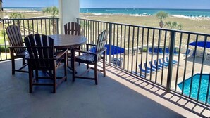 A balcony with a view of the beach and ocean, featuring a table and chairs, a pool, and umbrellas.