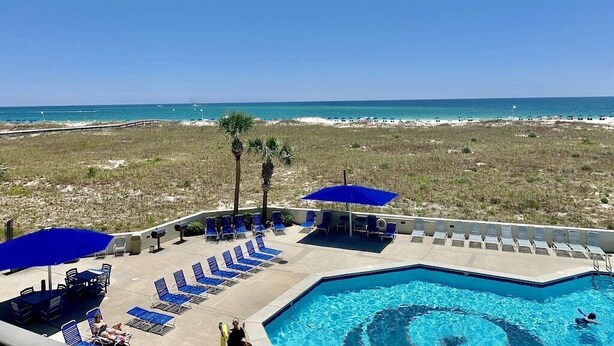 A pool area with blue lounge chairs and umbrellas, a clear view of the ocean, and a sandy beach in the background.