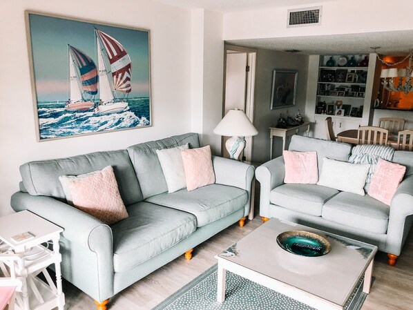 A living room with two light grey sofas, a small white coffee table, and a painting of sailboats on the wall. - Ocean One 306 - Oceanside prime beach location, walk to shops,dining, remodeled (Hilton Head Island)