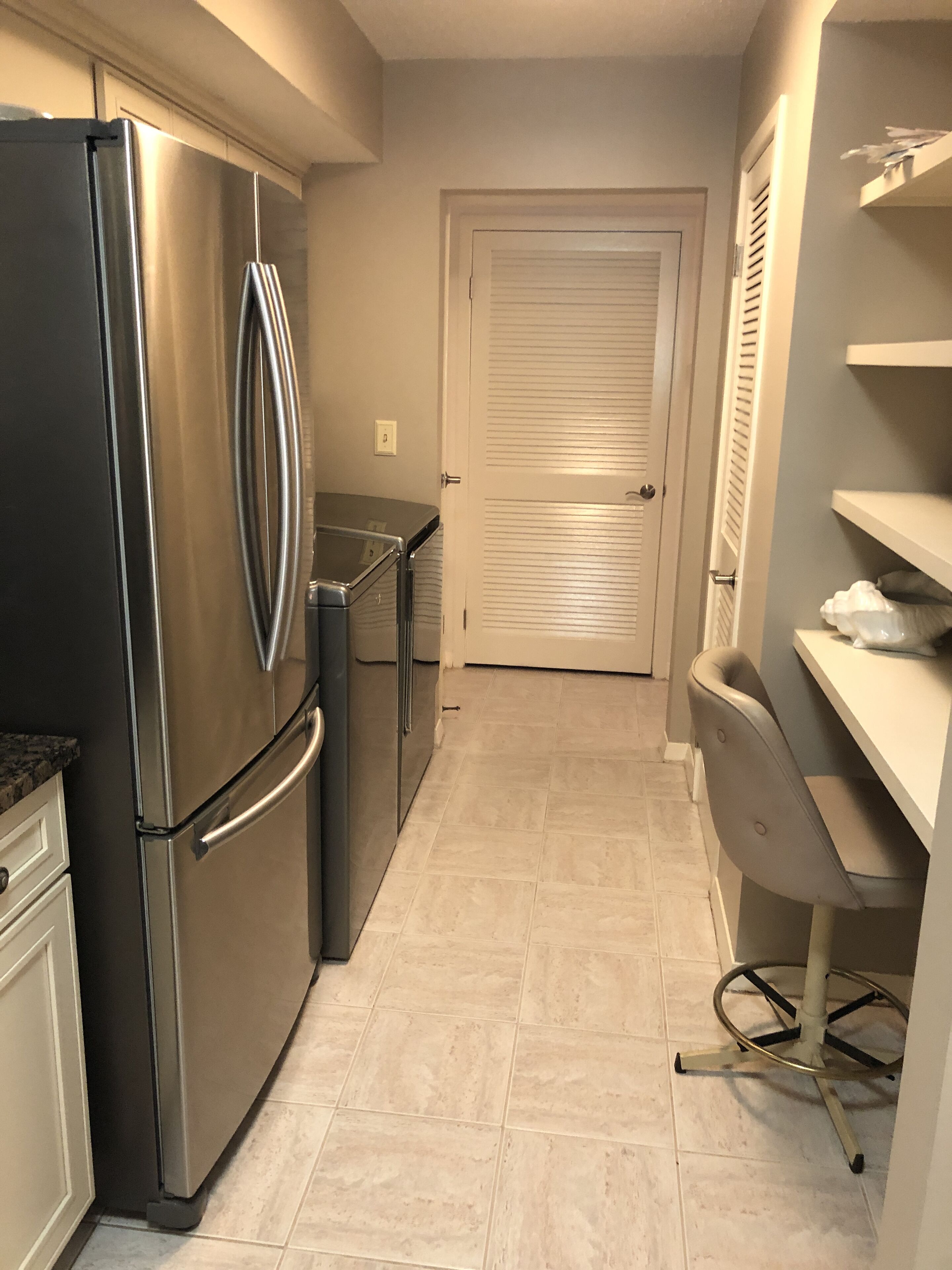 Cocina estrecha con electrodomésticos de acero inoxidable, una puerta blanca y una pequeña zona de trabajo con una silla y un escritorio.