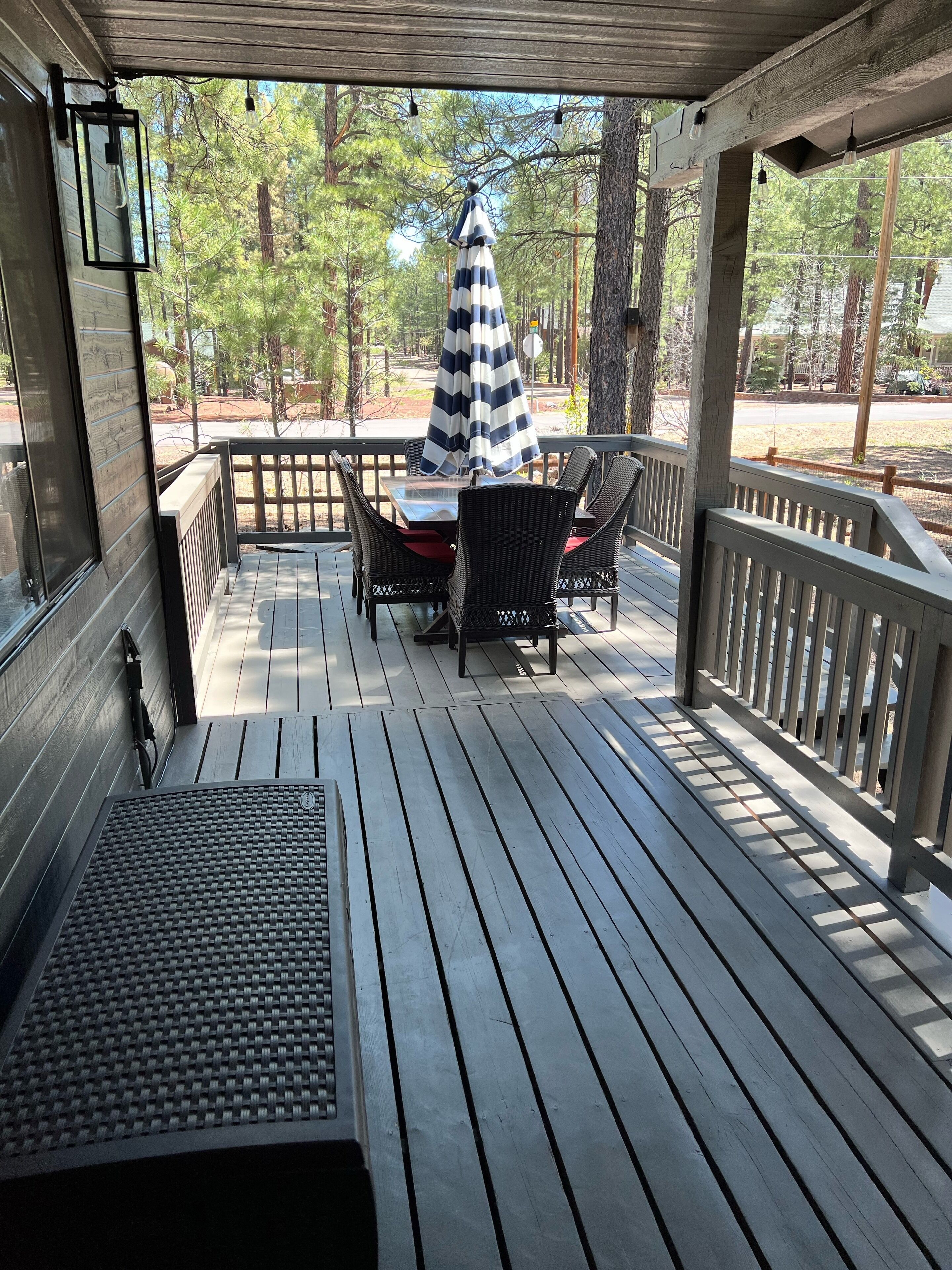A wooden deck with a table and chairs, a covered area with a striped umbrella, and a view of trees and a road.