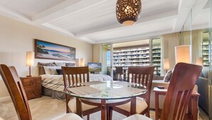 Une chambre d’hôtel avec une table à manger au plateau en verre, des chaises en bois, un lit donnant sur la mer, un tableau au mur et un balcon avec vue sur la ville.