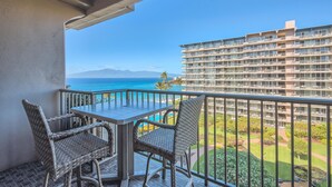 Un balcon avec des fauteuils en osier et une table, d’où l’on peut admirer un grand bâtiment et un ciel bleu dégagé.
