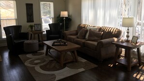 A living room with a sofa, two armchairs, a coffee table, and a side table with a lamp. There are two windows with blinds, a plant, and a welcome sign. - Estate Living! Professionally Cleaned, Self Check In - Sleeps 12 (Mt Pleasant)