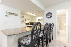 A kitchen with a granite countertop, four black chairs with patterned cushions, a wall clock, and a 'Happy Hour' sign.