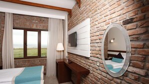 Habitación de hotel con pared de ladrillo, un ventanal con cortinas, cama con ropa de cama azul, escritorio con lámpara y un espejo que refleja la habitación.