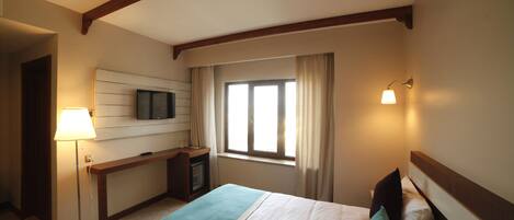 A hotel room with a bed, a desk, a TV, a window with curtains, and a lamp.