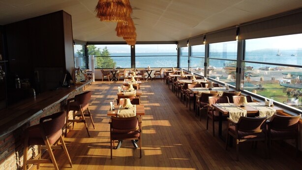 Un restaurante con suelo de madera, mesas y sillas, con amplios ventanales que dan al mar y a los barcos.