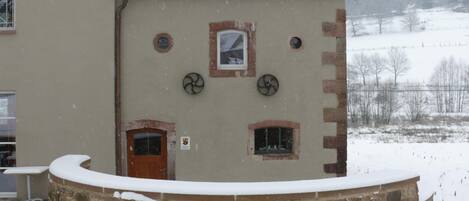 Un edificio con techo nevado y pared de piedra en primer plano. Se ven dos ventanas y una puerta.