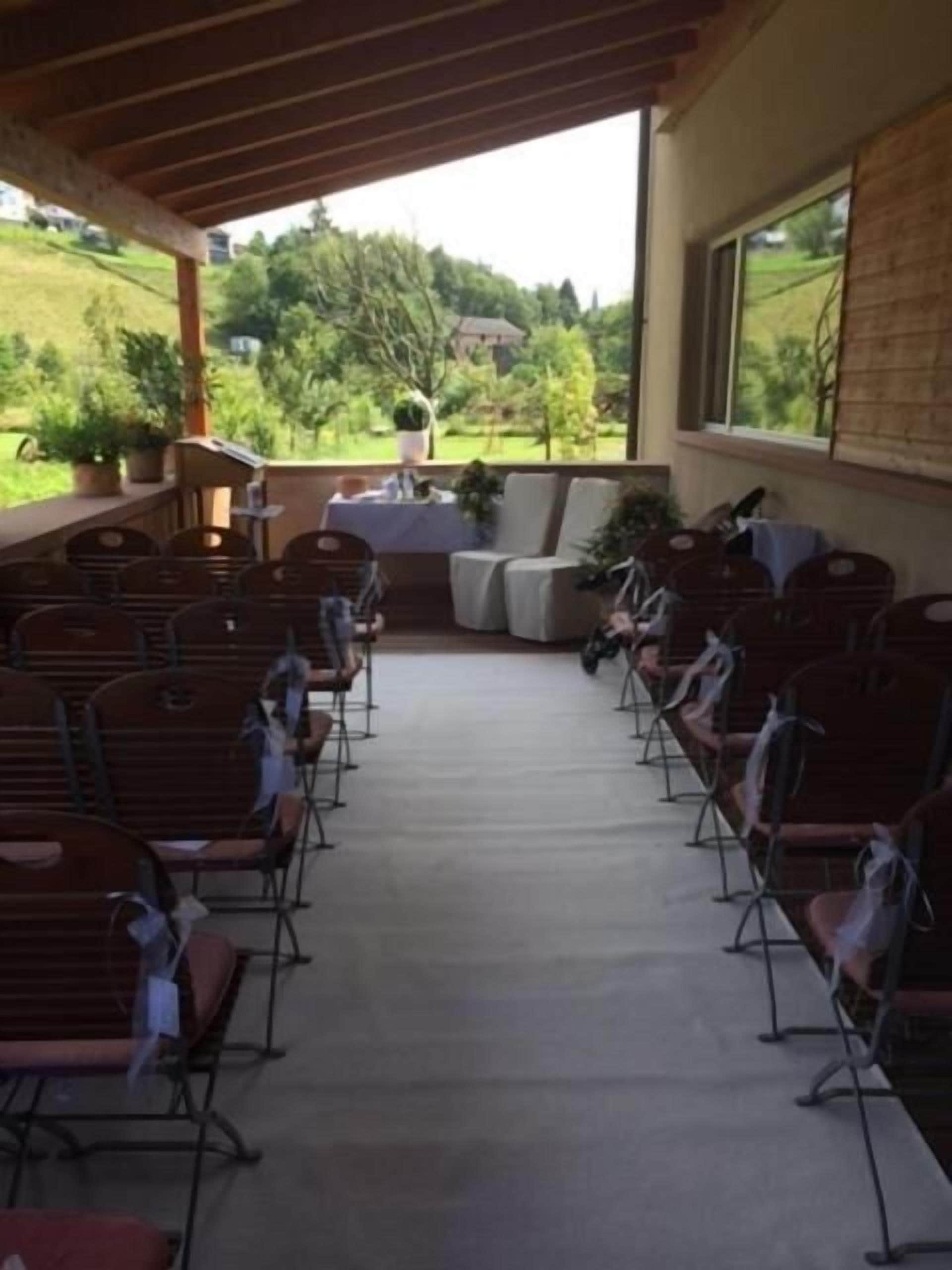 An outdoor seating area with rows of chairs and tables, a covered section with a table and chairs, and a view of greenery and buildings in the background.