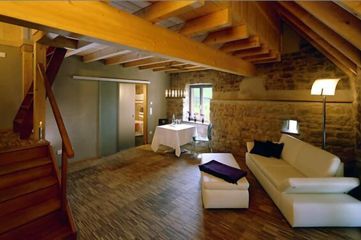 Ein geräumiger Lofts mit freiliegenden Holzbalken, einer Steinwand, einem weißen Sofa und einem Essbereich mit Tisch und Stühlen.