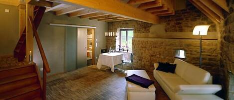 A spacious loft with exposed wooden beams, a stone wall, a white sofa, and a dining area with a table and chairs.