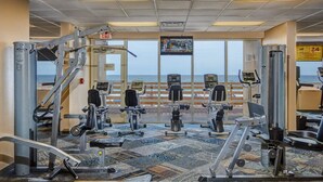 Ett gym med olika träningsredskap, ett stort fönster med utsikt över havet och en skylt som visar att det är öppet dygnet runt.