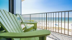 A balcony with a view of the beach, featuring green Adirondack chairs and a metal railing.