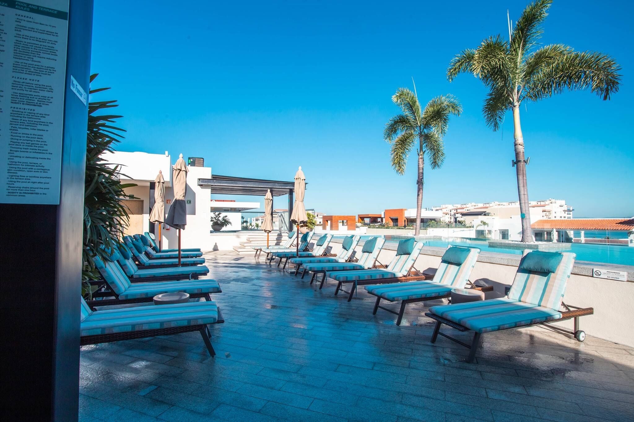 A poolside area with lounge chairs, palm trees, and a clear blue sky.