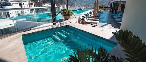 A rooftop infinity pool with clear blue water, surrounded by lounge chairs and umbrellas, overlooking a mountainous landscape.
