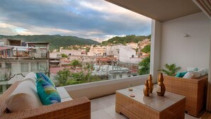 Un balcón con mobiliario de mimbre, una mesa de vidrio y vistas a un paisaje urbano con edificios y colinas.