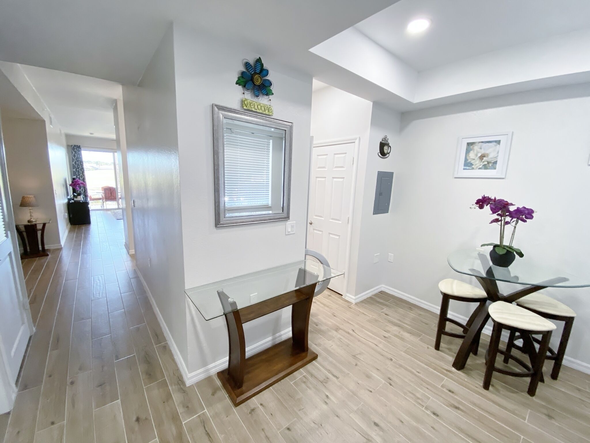 A modern dining area with a glass table and wooden chairs, a welcome sign, a window with blinds, and a door leading to another room.