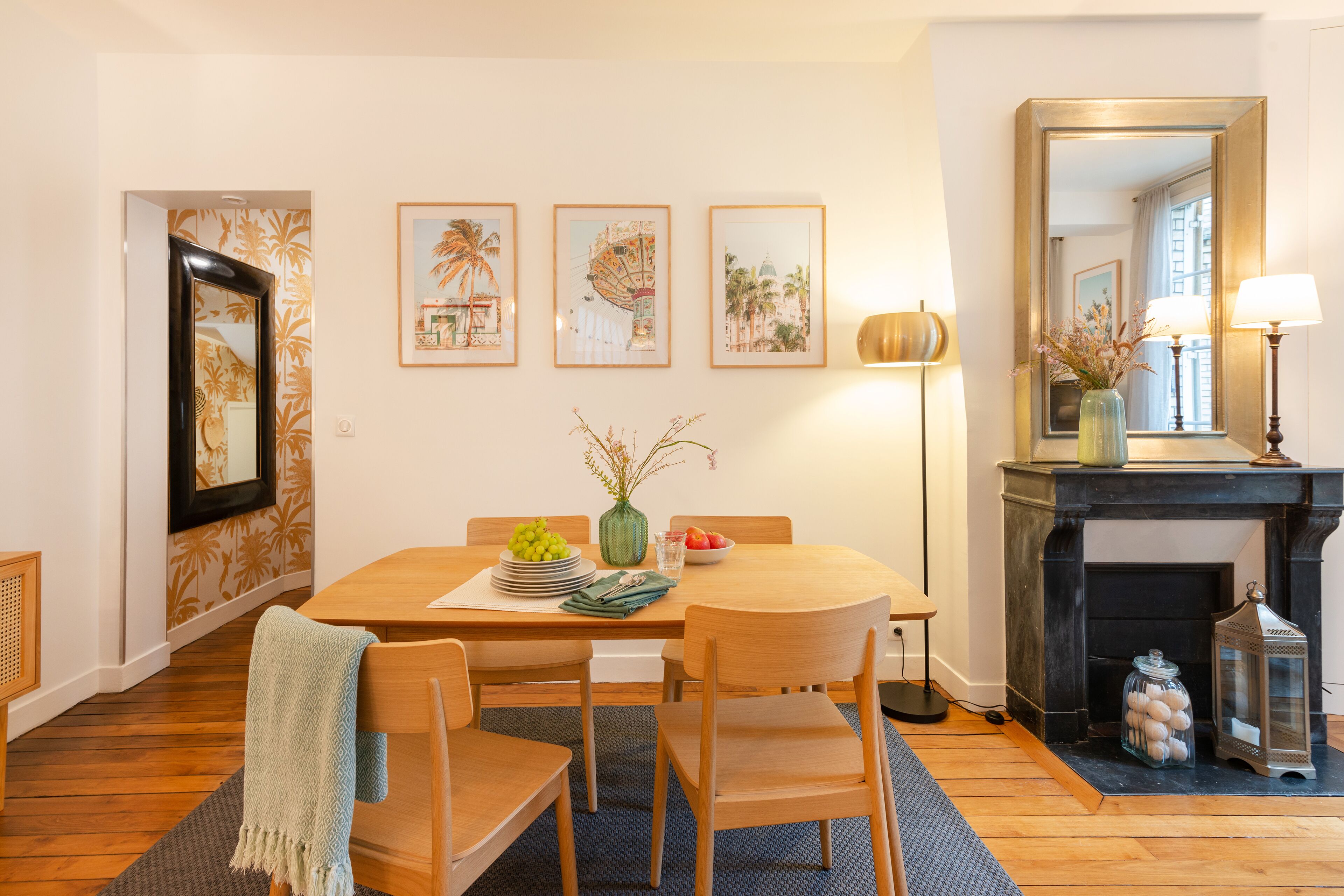 A dining area with a wooden table, chairs, and a vase with flowers. There are framed pictures on the wall and a mirror above the table.