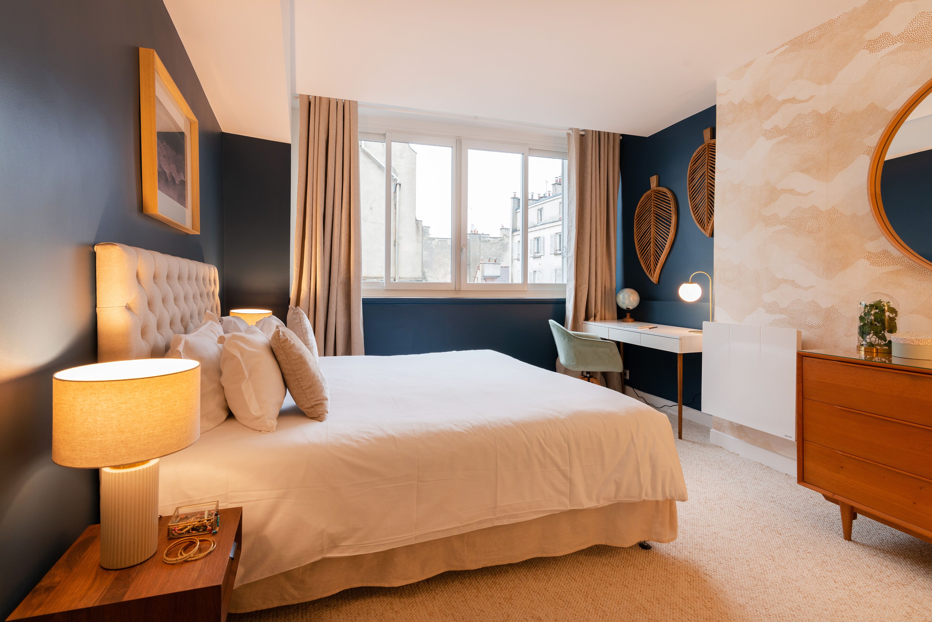 A bedroom with a bed, bedside lamp, desk, chair, dresser, and a window with curtains.