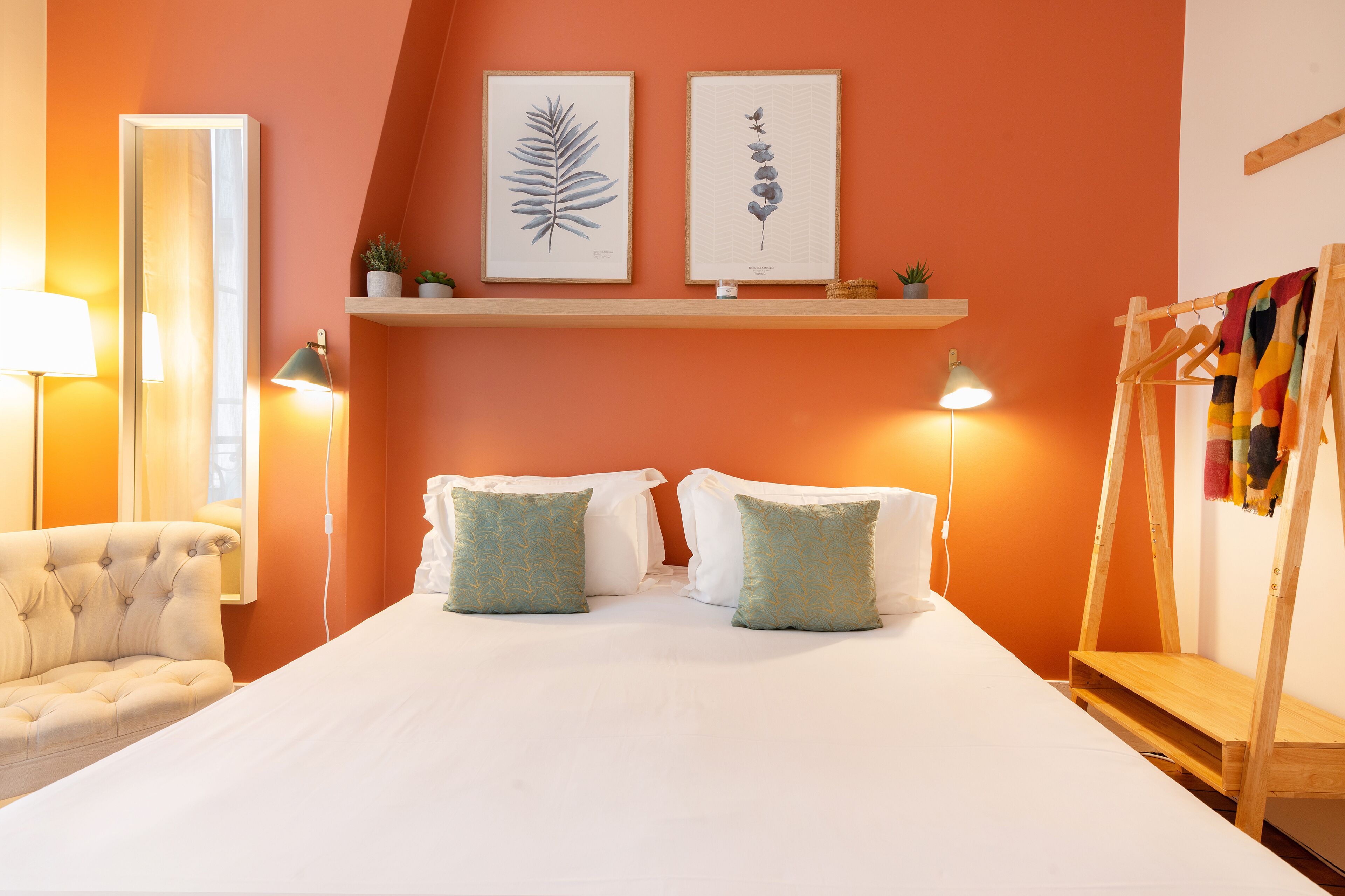 Una camera da letto con un letto, due cuscini, una sedia, un comodino, una lampada, uno specchio e due mensole a parete con piante.