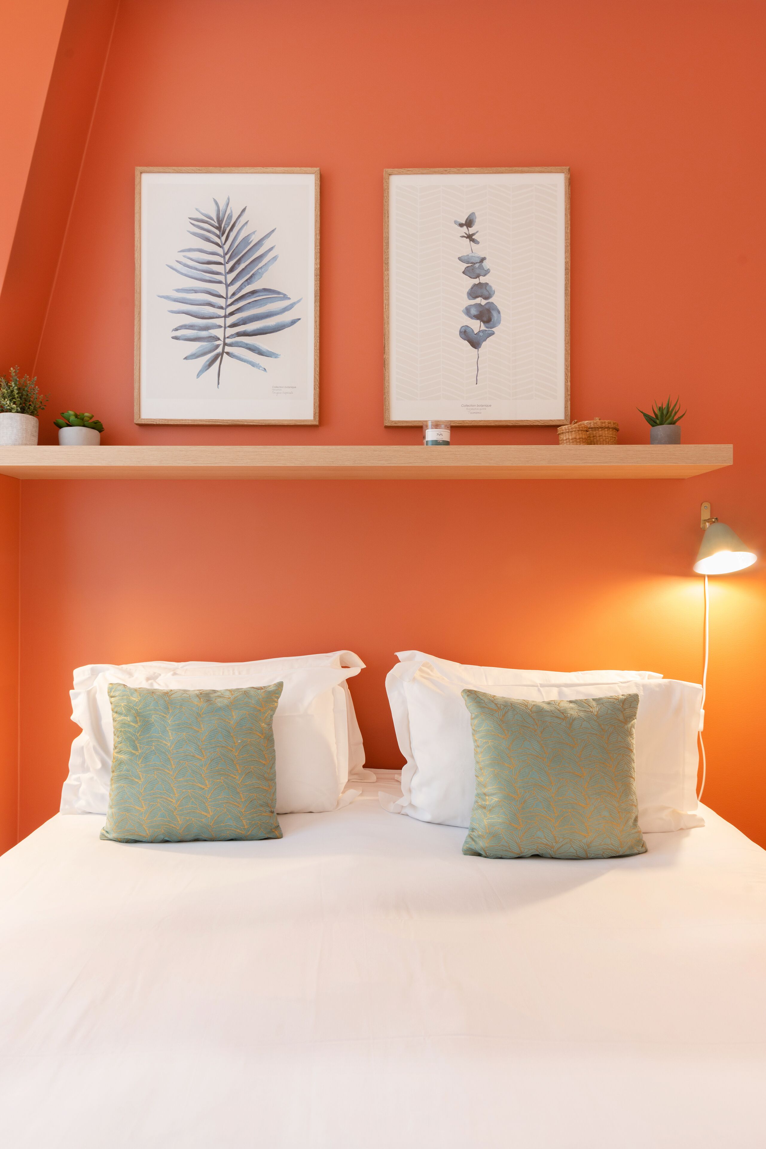 Une chambre à coucher avec un lit, deux oreillers, une table de chevet, une lampe et deux reproductions botaniques encadrées accrochées au mur.