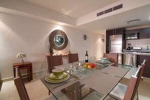A modern dining area with a glass table, wooden chairs, and a kitchen in the background.