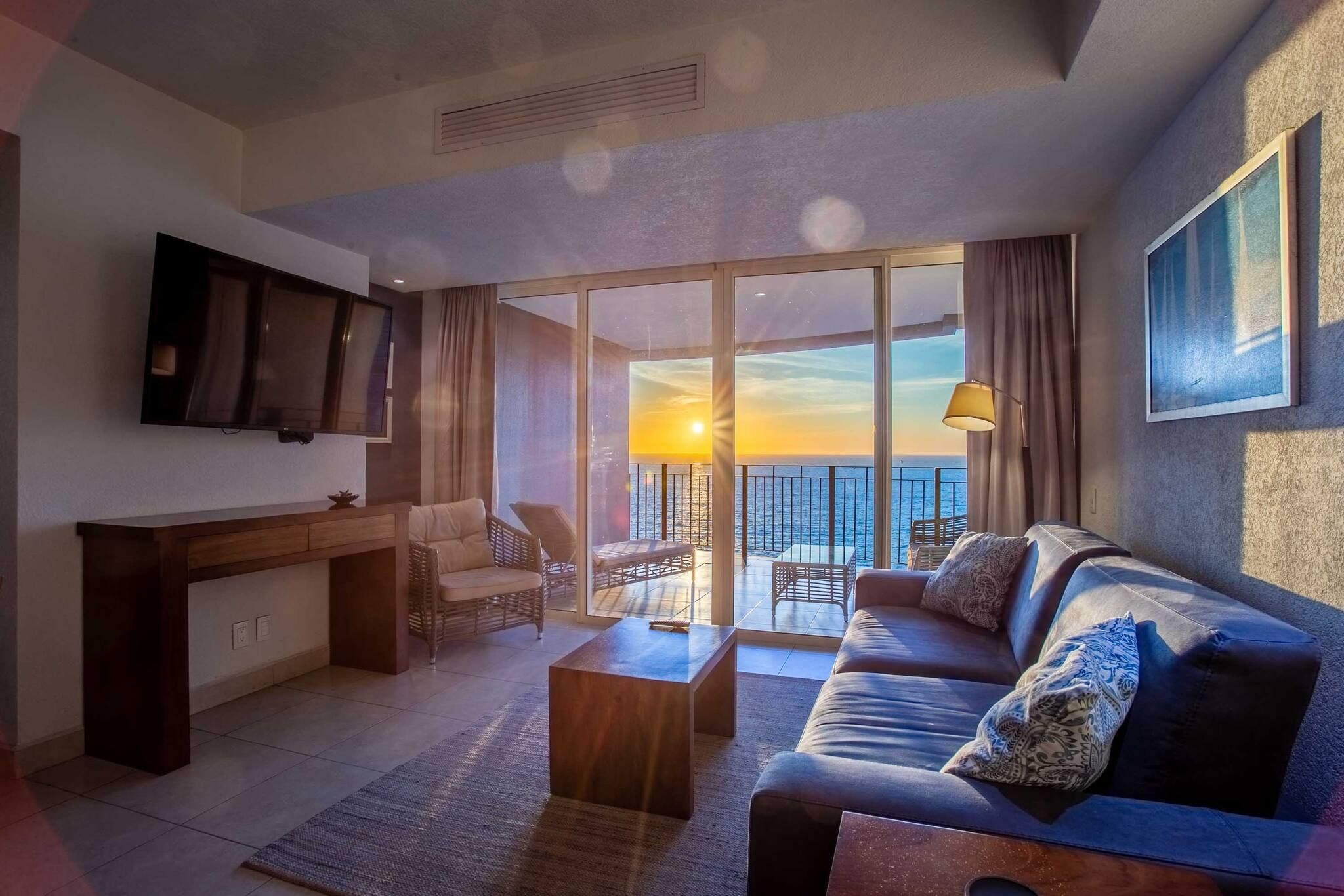 Un salon moderne avec un canapé, une table basse et un téléviseur à écran plat. Il y a un balcon d’où l’on peut admirer le coucher du soleil.