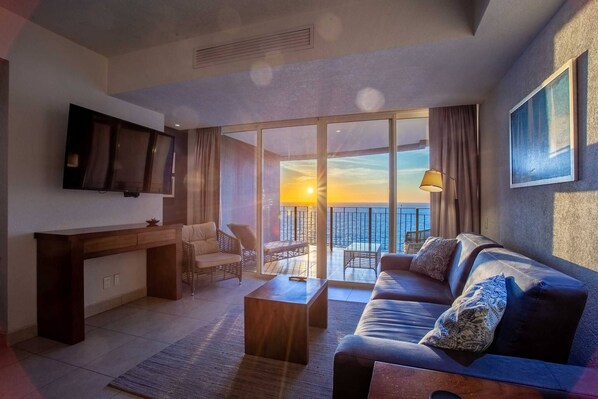A modern living room with a sofa, coffee table, and a flat-screen TV. There is a balcony with a view of the sunset.
