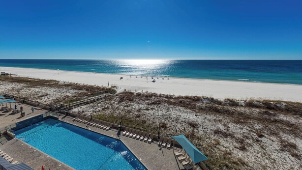 A beachside pool area with a clear view of the ocean, a sandy beach, and a small building with a blue roof. - Phoenix X - 6th Floor - 3 Bedroom (Orange Beach)