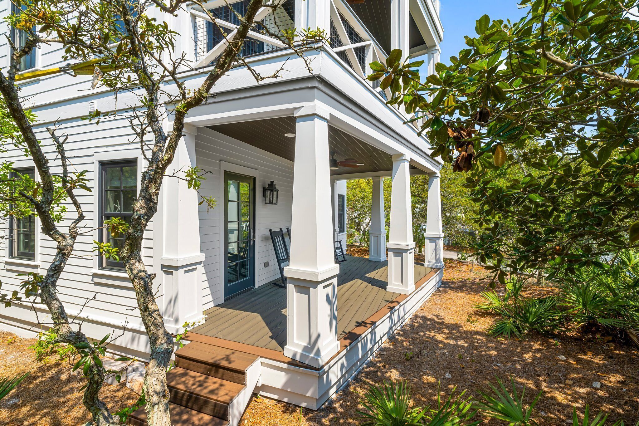 A white house with a wooden porch and columns, surrounded by trees and shrubs.