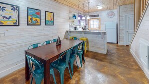Een eetgedeelte met een houten tafel en teal stoelen, een keuken met een koelkast en een 'HAPPY HOUR' bord, en een houten plafond.