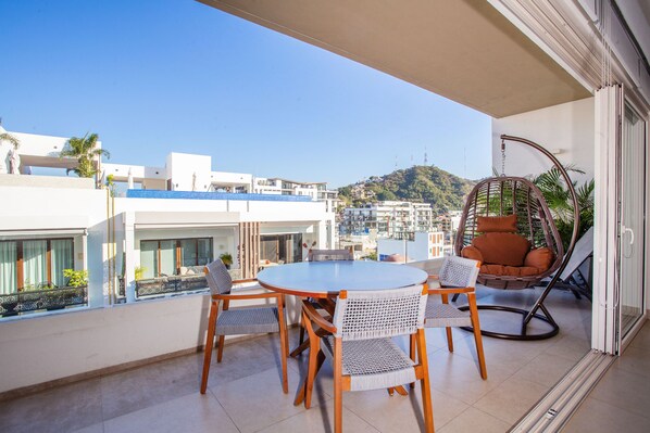 A balcony with a round table and chairs, a wicker chair, and a view of buildings and hills.