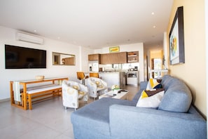 A modern living room with a grey sofa, a wooden bench, and two patterned armchairs. A kitchen with wooden cabinets and a dining area with a table and chairs is visible in the background. - Top Floor Condo with City View, Rooftop Pool, Gym in Old Town (Puerto Vallarta)