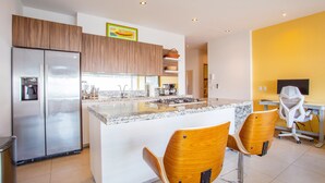 A modern kitchen with a stainless steel refrigerator, wooden cabinets, a marble countertop, and two wooden bar stools. - Top Floor Condo with City View, Rooftop Pool, Gym in Old Town (Puerto Vallarta)