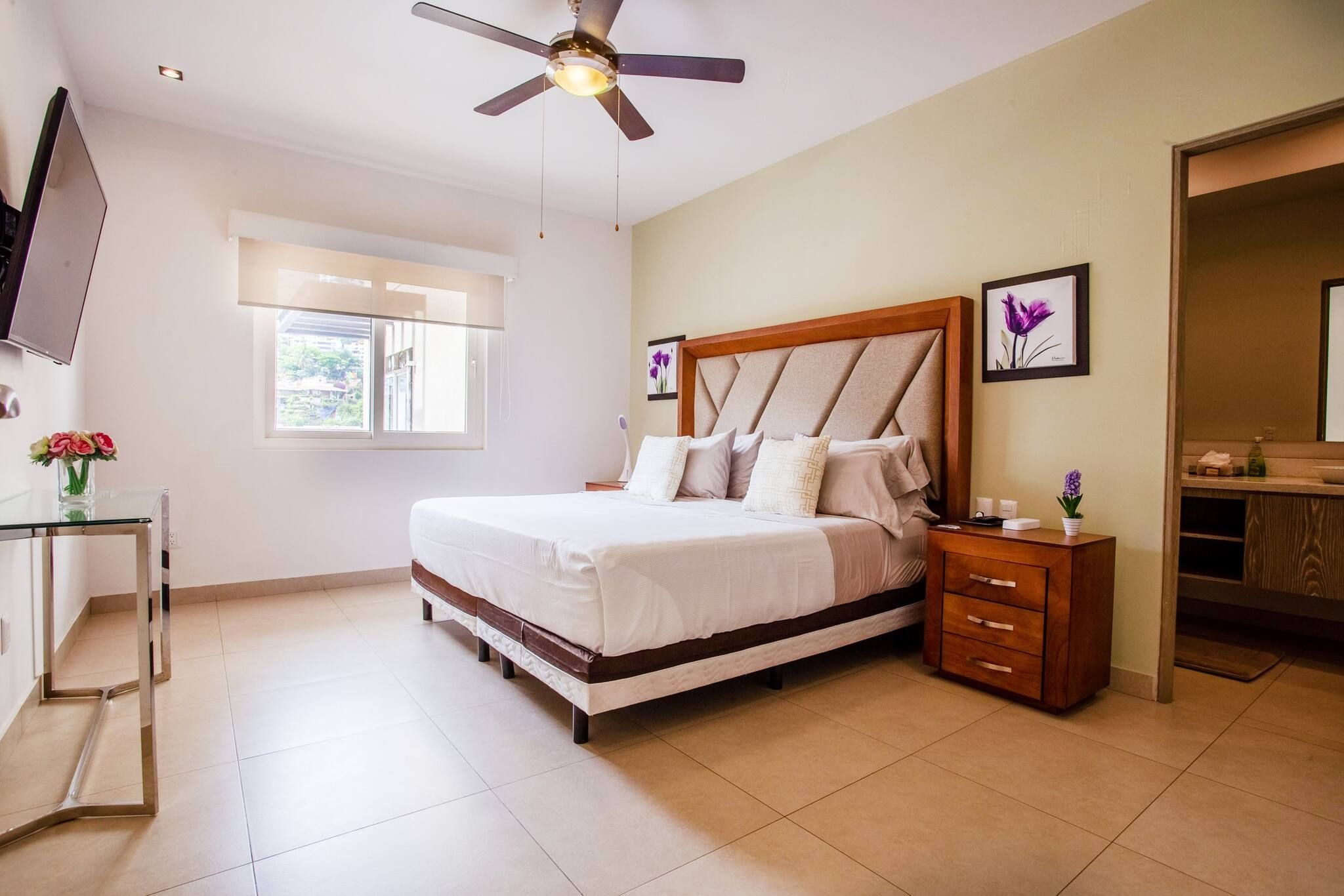 A bedroom with a bed, a bedside table, a nightstand, a ceiling fan, a window, and a television.